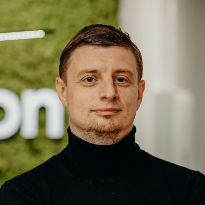 A man standing in the office in front of the Kellton sign, wearing a black turtleneck