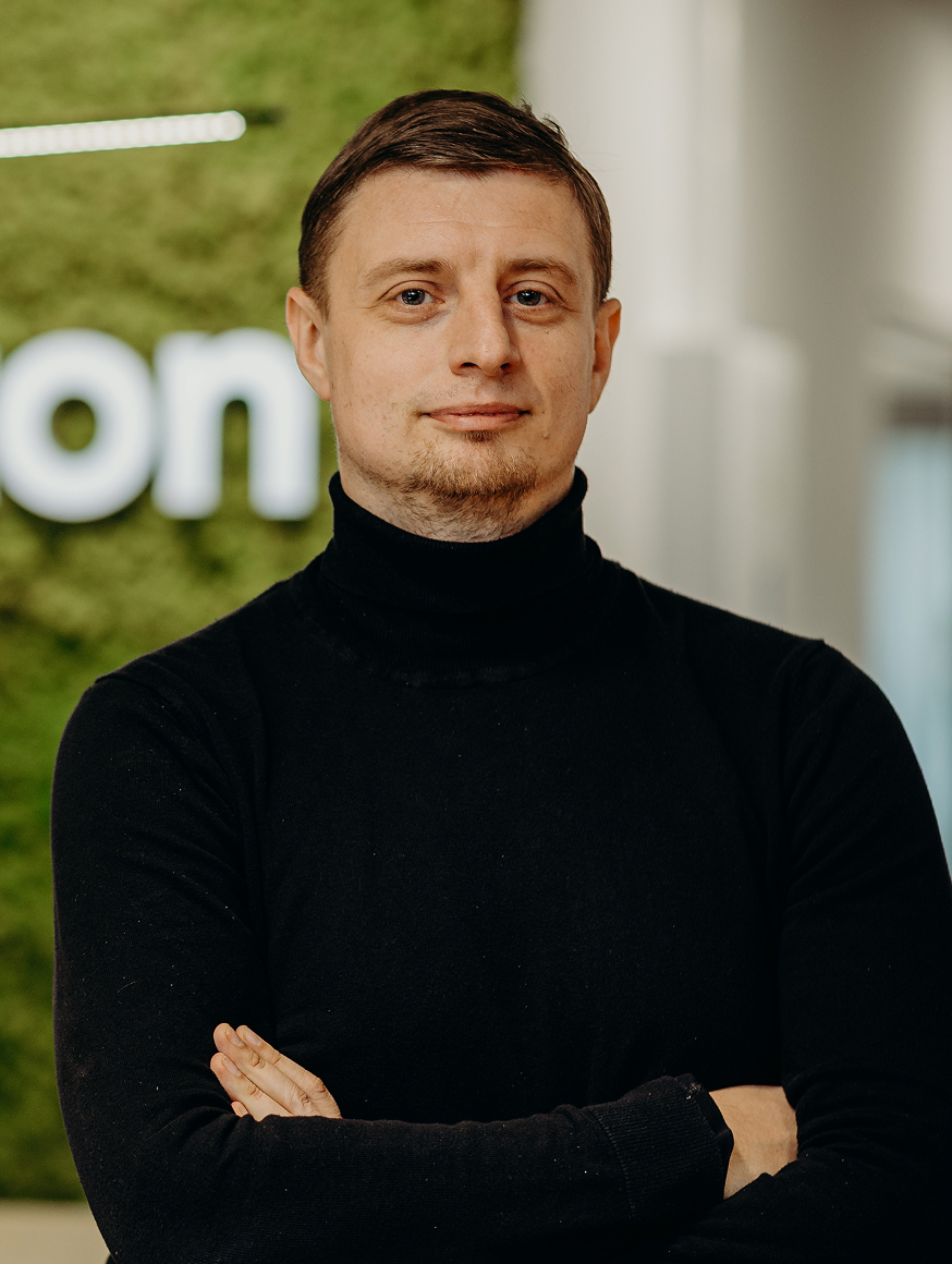 A man standing in the office in front of the Kellton sign, wearing a black turtleneck