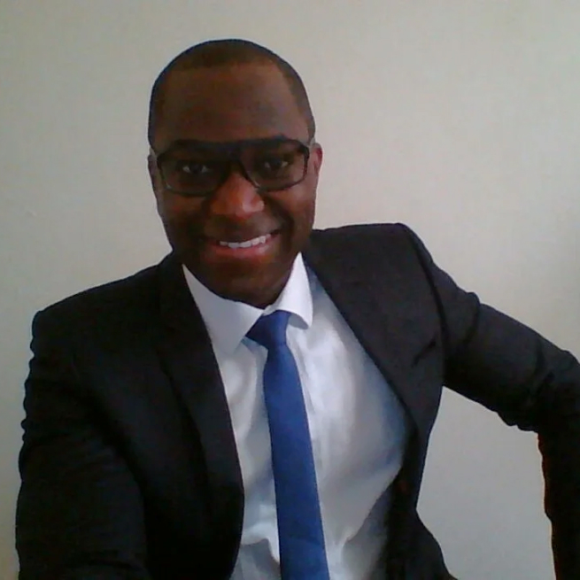A man wearing glasses, a dark suit, white shirt, and blue tie is smiling at the camera. He is seated and leaning slightly forward with one arm resting on the side, against a plain light-colored background.