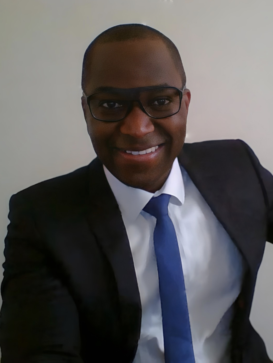 A man wearing glasses, a dark suit, white shirt, and blue tie is smiling at the camera. He is seated and leaning slightly forward with one arm resting on the side, against a plain light-colored background.