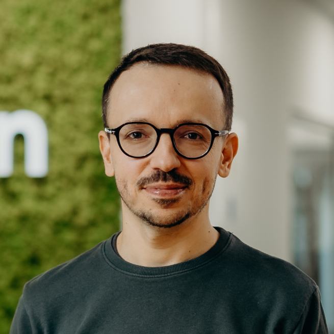A man standing in the office in front of the Kellton sign, wearing a green shirt and glasses.