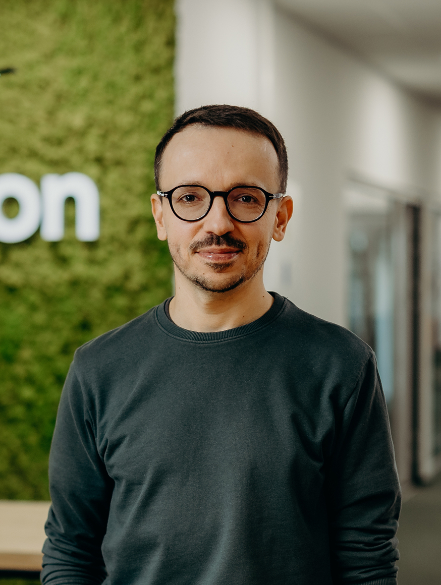 A man standing in the office in front of the Kellton sign, wearing a green shirt and glasses.