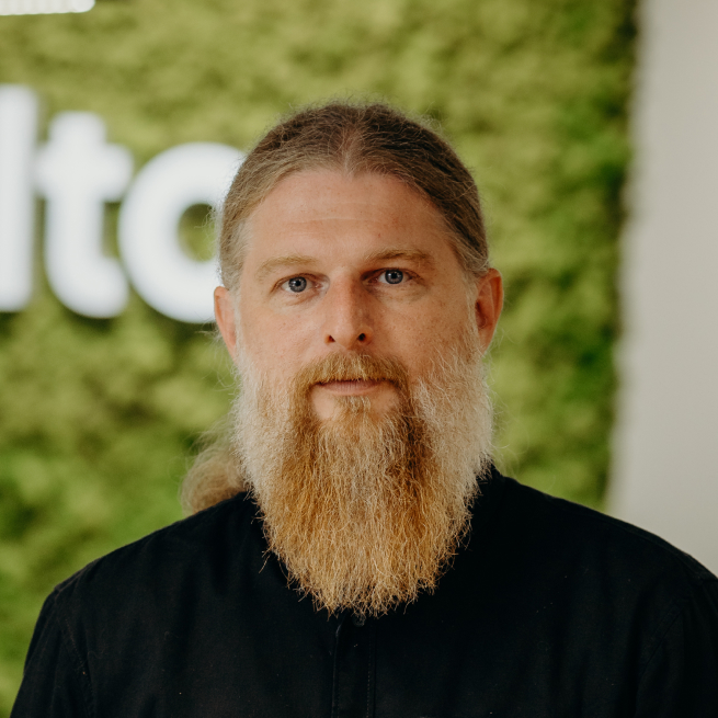 A man with a long beard standing in the office in front of the Kellton sign, wearing a black shirt.