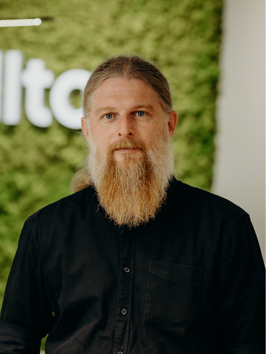 A man with a long beard standing in the office in front of the Kellton sign, wearing a black shirt.