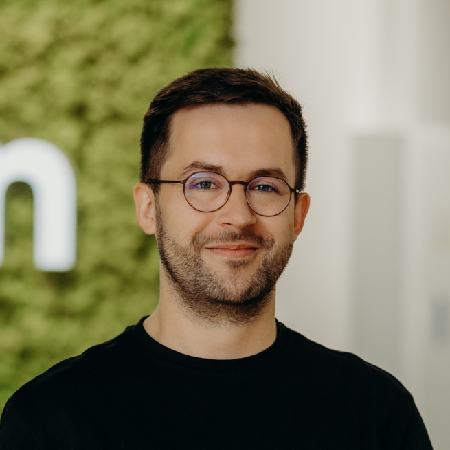 A man standing in the office in front of the Kellton sign, wearing a black shirt and glasses.