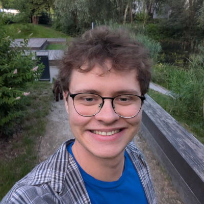 Smiling man wearing glasses and a casual shirt, standing outdoors with a green park background.
