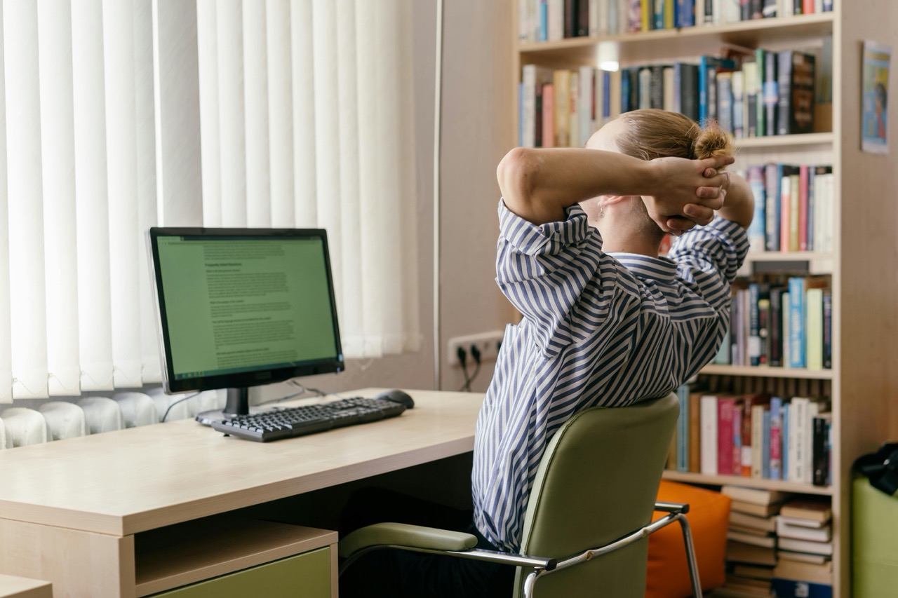 Person in striped shirt stretching at desk with computer, bookshelf in background.