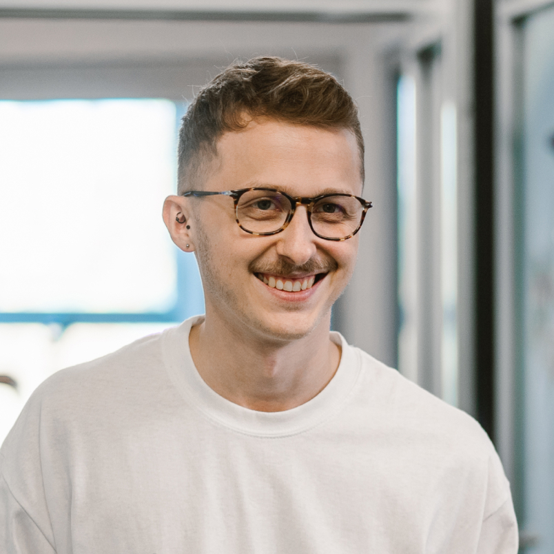 A man standing in the office and smiling, wearing glasses and a white shirt
