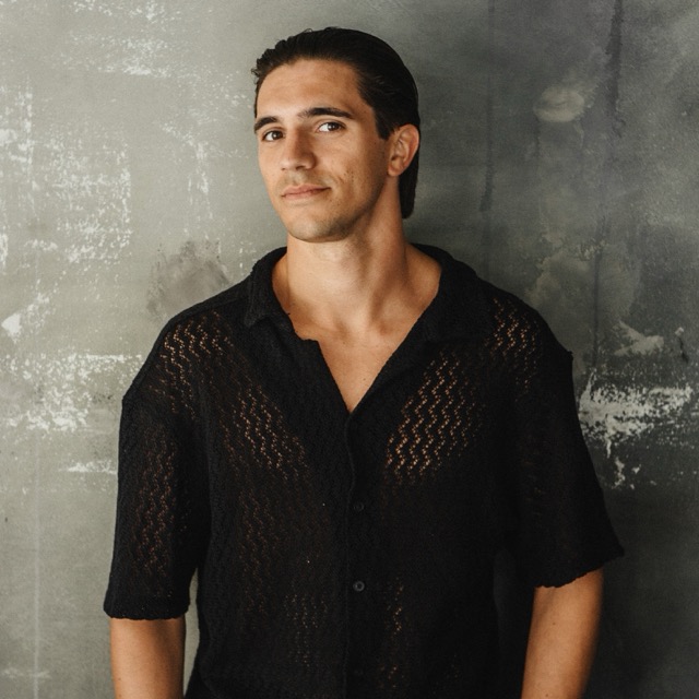 A man in a black patterned shirt standing against a textured wall.