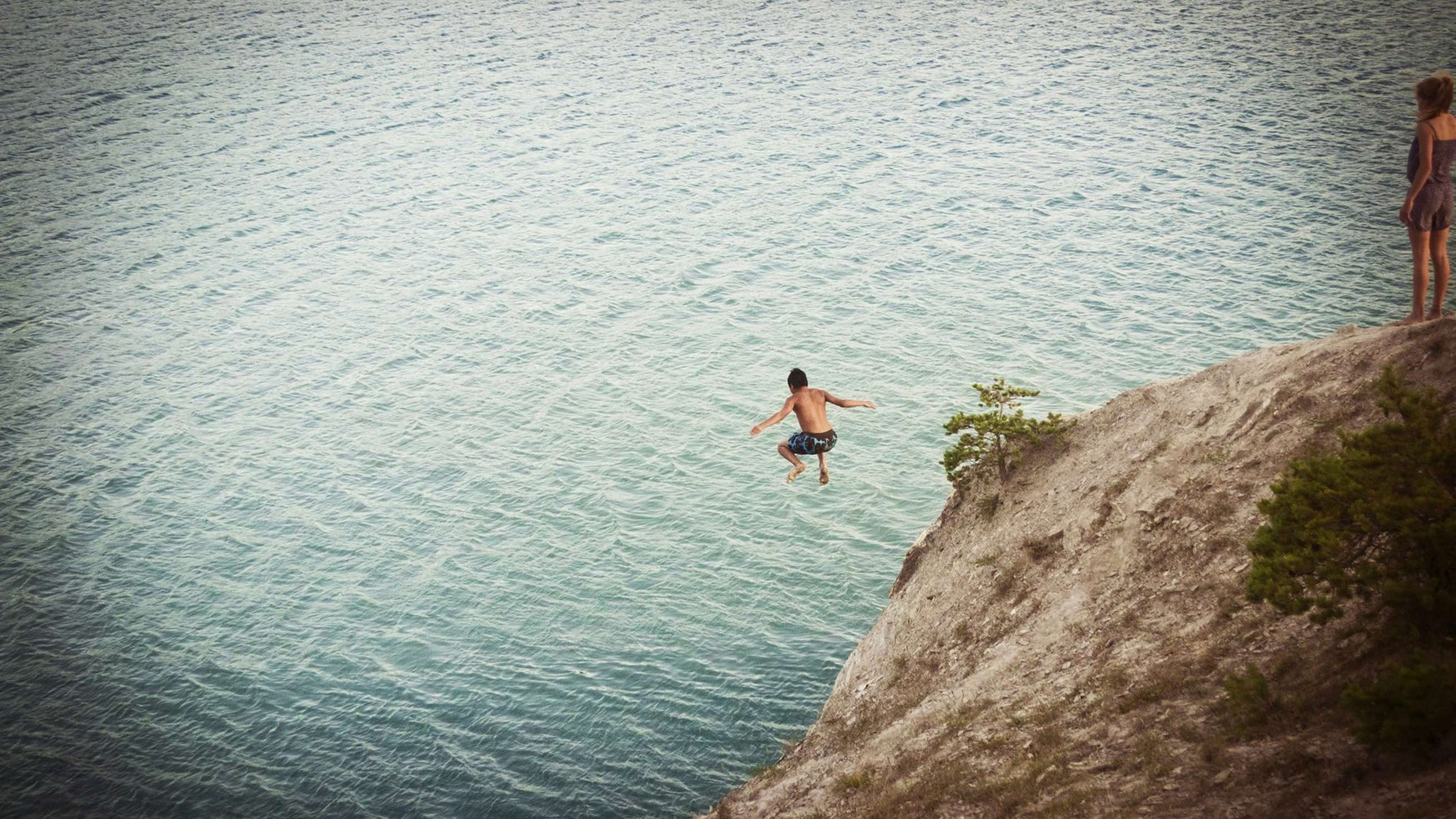 Person cliff diving into blue water while another watches