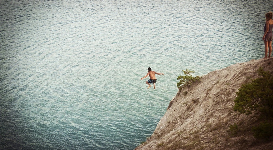 Person cliff diving into blue water while another watches