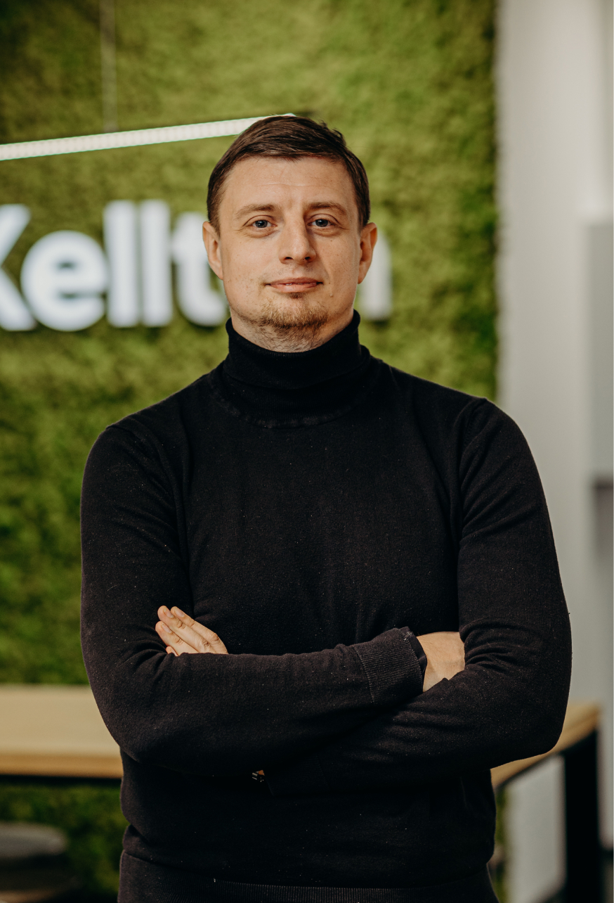 A man standing in front of the Kellton sign in the office, wearing a black turtleneck