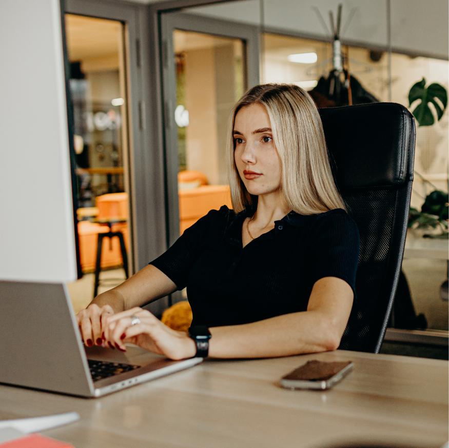 A person with long hair, focused on their laptop, seated at a desk in an office environment.