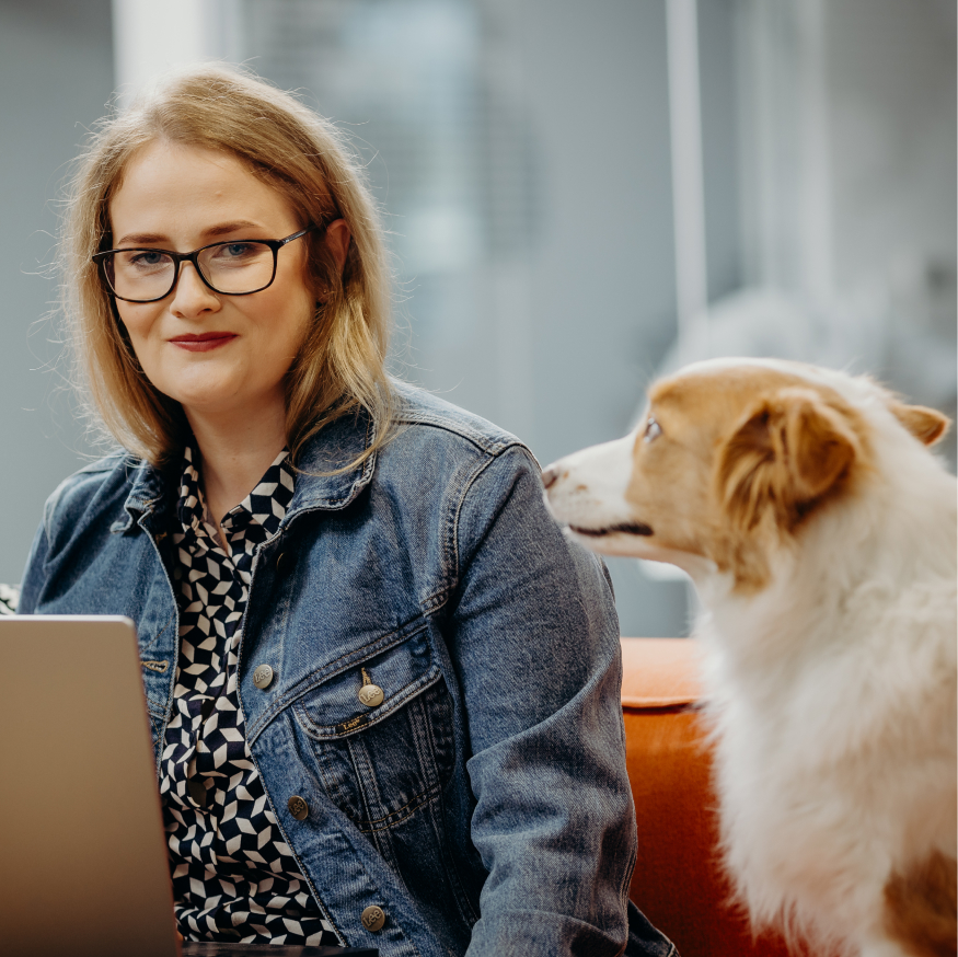 A person in glasses and a denim jacket working on a laptop, with a dog sitting beside them.