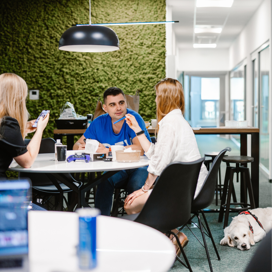 Three people sitting around a table in a well-lit office with a green wall, engaged in conversation.