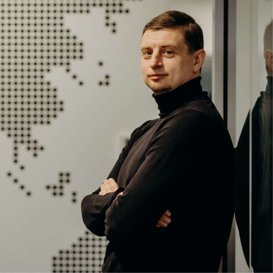 A man in a black turtleneck stands with his arms crossed, looking at the camera. He has short brown hair. The background features a wall with a dotted world map design, and a glass surface reflects part of his image.
